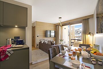 Cozy, modern ski apartment with breakfast on the dining table, open-plan living, and a balcony boasting snowy mountain views.