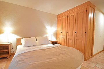 A warm-toned bedroom features a bed with white pillows, two lighted nightstands, a large wooden built-in wardrobe, and terracotta-colored tiled flooring.