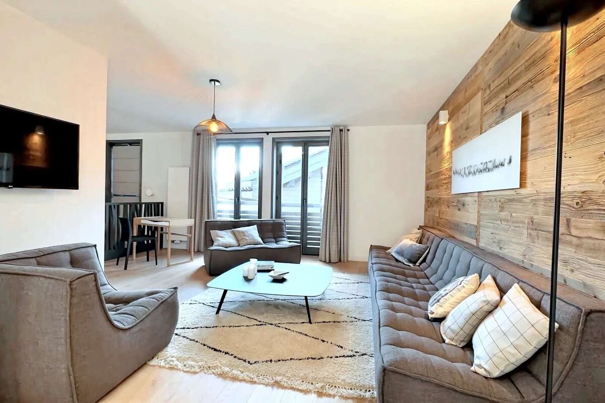 Cozy, modern living room featuring gray tufted seating, a rustic wood accent wall, a geometric rug, and large windows leading to a balcony.