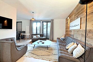 Cozy, modern living room featuring gray tufted seating, a rustic wood accent wall, a geometric rug, and large windows leading to a balcony.