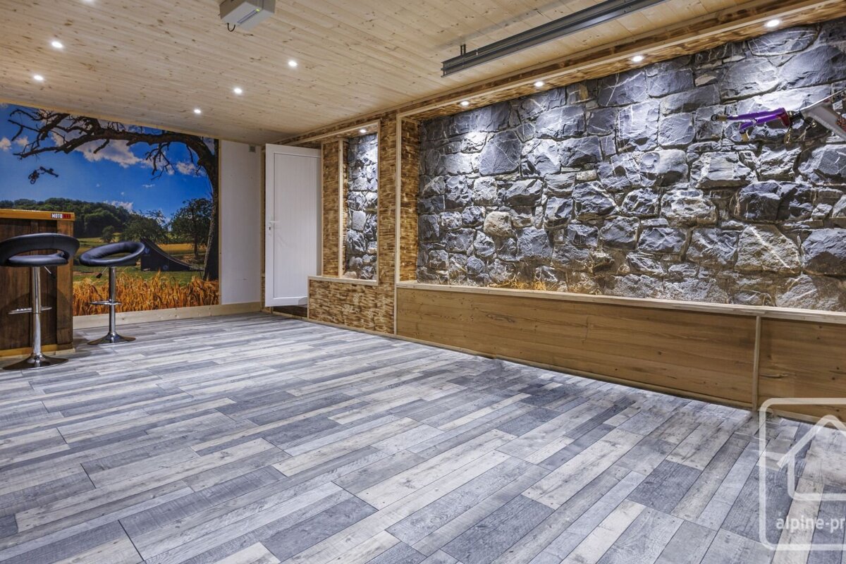 A contemporary rustic room boasts a stone accent wall, wood ceiling, scenic mural, a bar with stools, and grey plank flooring.