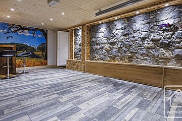 A contemporary rustic room boasts a stone accent wall, wood ceiling, scenic mural, a bar with stools, and grey plank flooring.