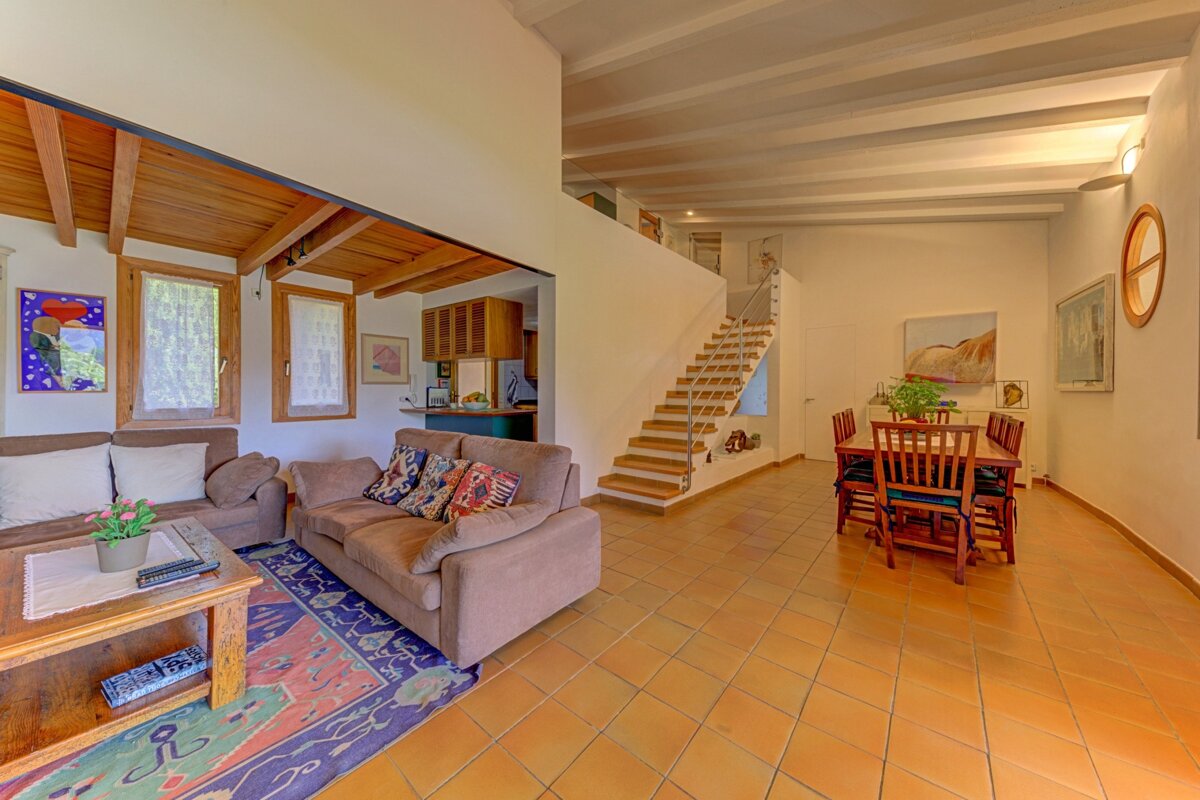 Spacious open-plan living room, dining area, and kitchen with tiled floors, a wooden staircase, cozy sofas, and colorful artwork.
