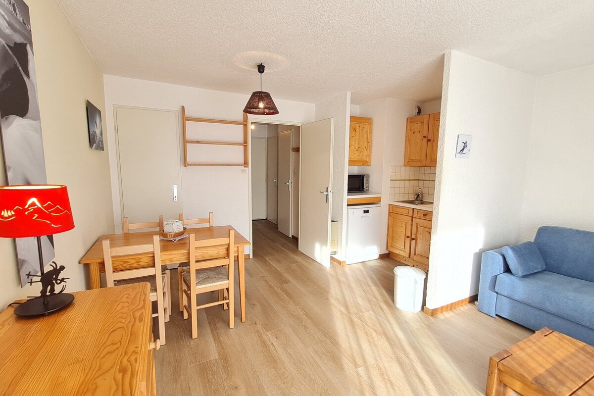 A bright, cozy studio apartment with light wood floors, a dining table, a small kitchen, and a blue sofa.