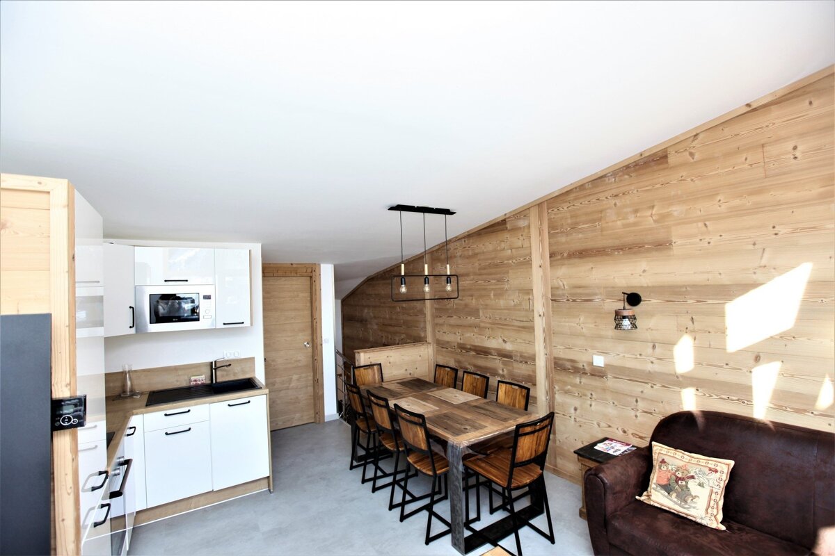 Modern open-plan space with a white kitchen, long wooden dining table and high chairs, and a brown sofa against a wood-paneled wall.