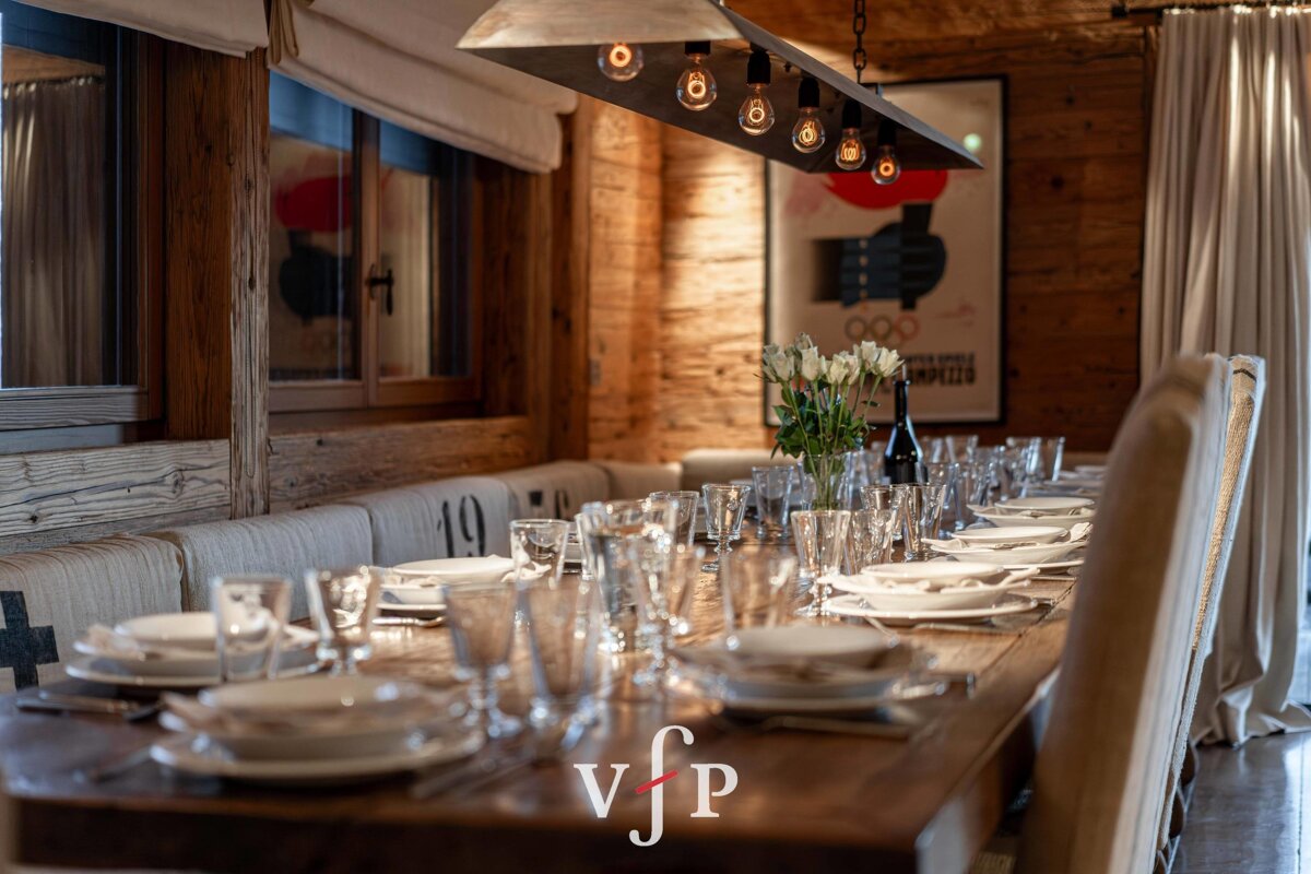 A cozy, rustic chalet dining room featuring a long wooden table elegantly set for a meal under warm overhead lighting.