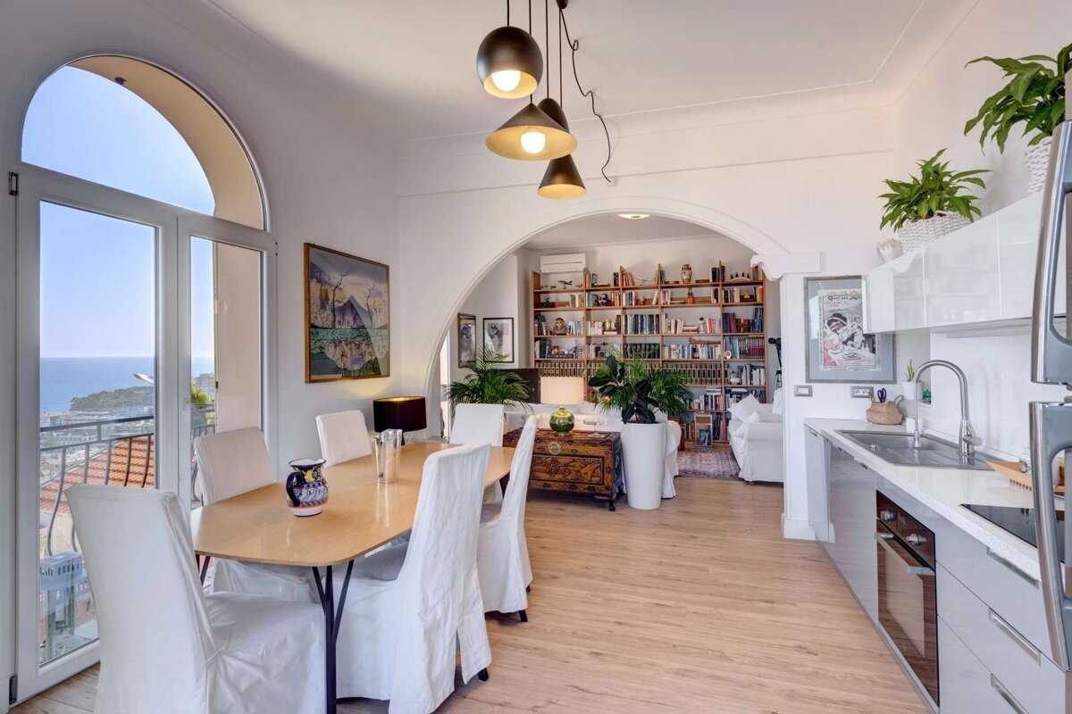 Bright open-plan dining, modern kitchen, and living room with a bookcase, all featuring a stunning sea view from an arched window.