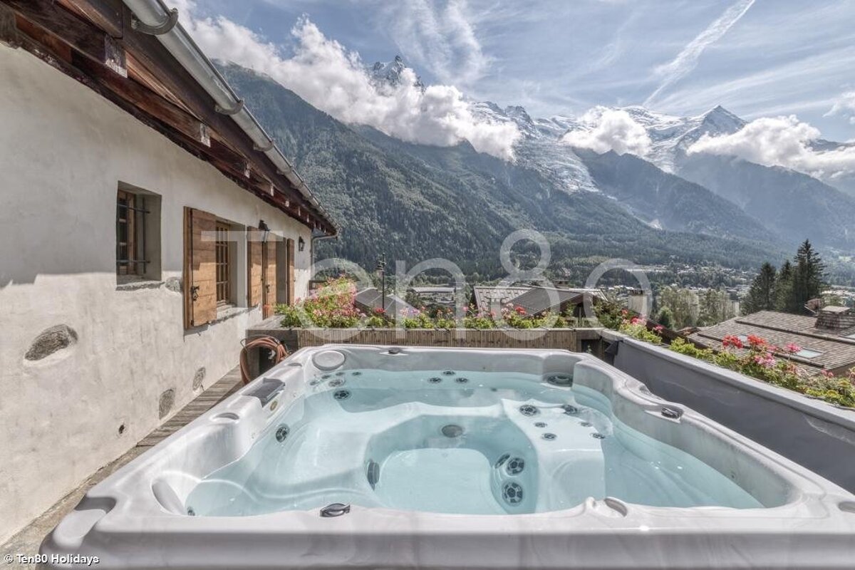 An outdoor hot tub on a chalet deck overlooking a stunning panorama of snow-capped mountains and a lush valley.