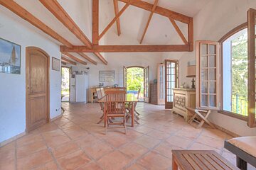 A spacious, sunlit room with exposed wooden beams, terracotta floor, dining table, and open doors to a lush garden view.