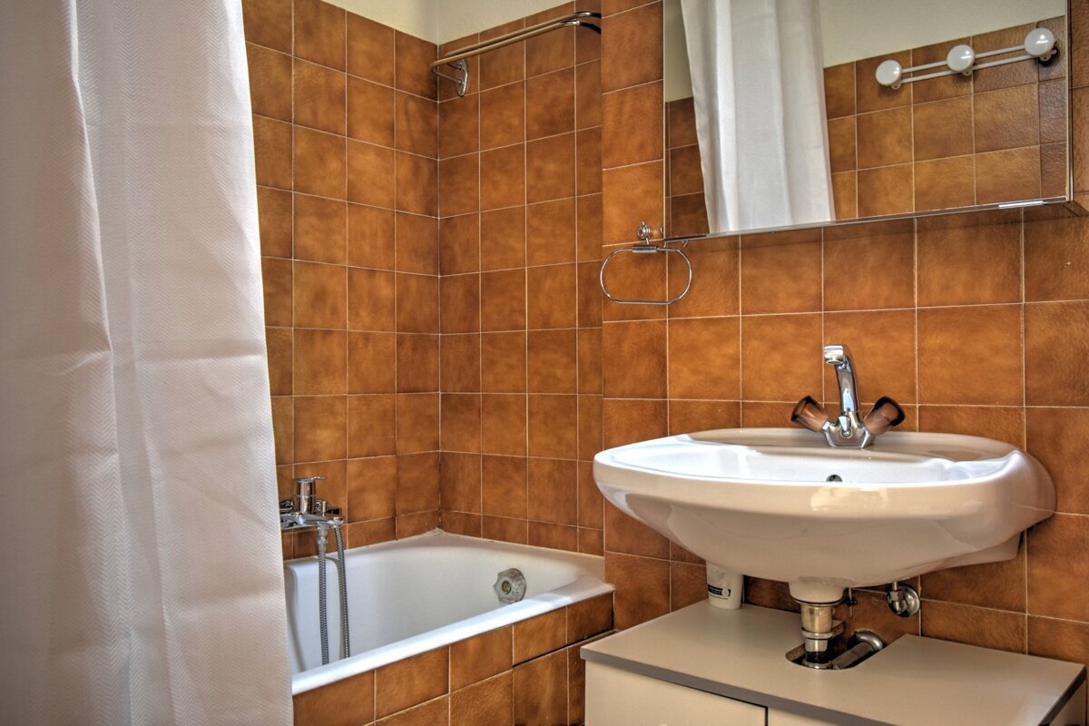 A bathroom with brown tiled walls, a white bathtub and shower curtain, and a sink with a mirror and lighting fixtures above it.