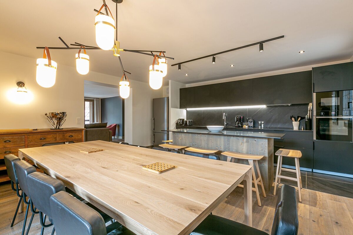 Bright, modern open-plan dining area with a large wooden table, black chairs, and an elegant kitchen featuring dark cabinets and a stylish island.