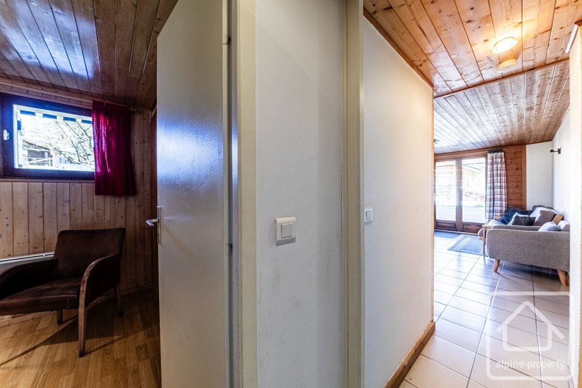 An interior shot shows a hallway dividing views into two rooms. Left: wood-paneled room with window and armchair. Right: living area with sofa, tiled floor, and glass doors. Both have wood ceilings.