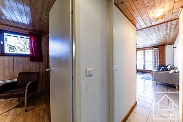 An interior shot shows a hallway dividing views into two rooms. Left: wood-paneled room with window and armchair. Right: living area with sofa, tiled floor, and glass doors. Both have wood ceilings.