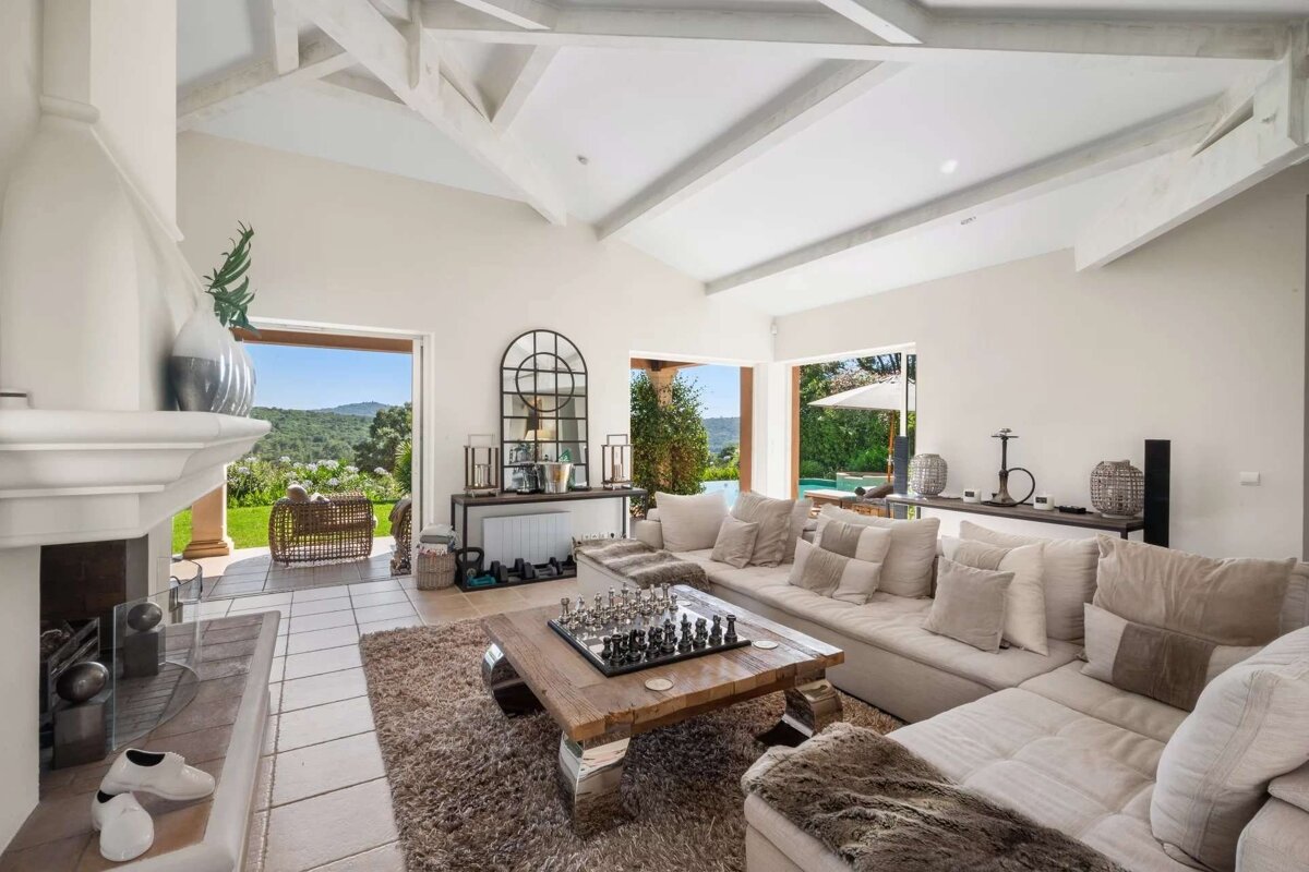 A bright, elegant living room with a huge beige sofa, chessboard coffee table, vaulted ceilings, and stunning views of a green landscape and outdoor pool.