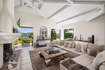 A bright, elegant living room with a huge beige sofa, chessboard coffee table, vaulted ceilings, and stunning views of a green landscape and outdoor pool.