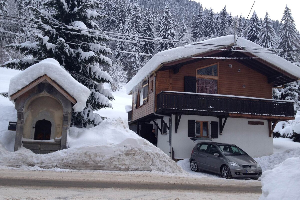 A snow-covered alpine chalet with a small shrine, a gray car parked, and a dense snowy forest in the background on a winter day.