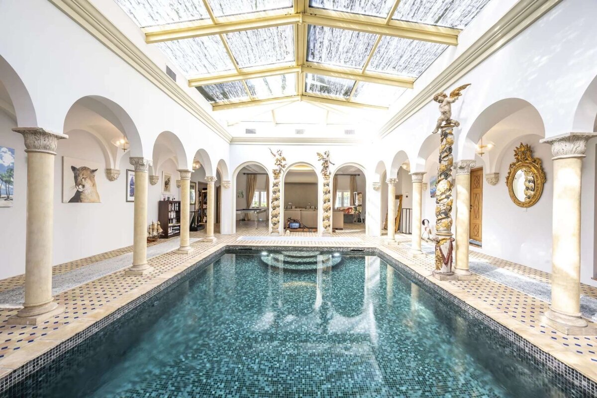 A luxurious indoor pool area features a glass skylight, surrounded by white arched colonnades, intricate tiling, and ornate classical decor.