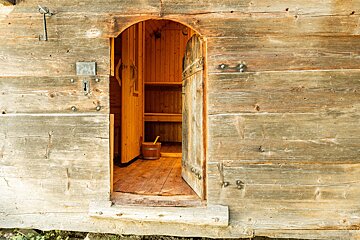 A rustic wooden building with an arched, open doorway revealing a warm sauna interior with benches and a bucket. An old key hangs on the exterior wall.