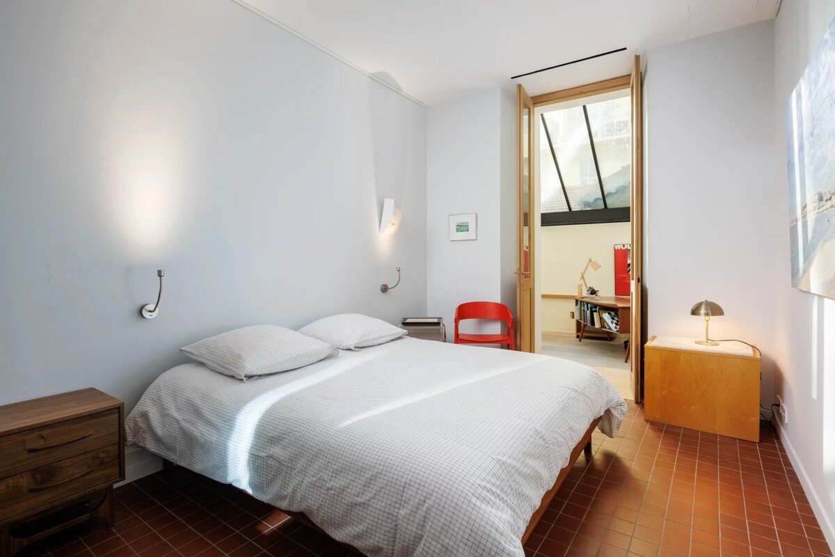 A bright, minimalist bedroom with a white bed on reddish-brown tiled floor. Features wooden furniture, light walls, and an open doorway to another room.