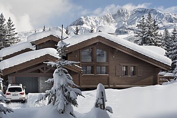 A cozy, snow-covered wooden chalet sits in a winter mountain wonderland, surrounded by snowy pines with a white car parked outside.
