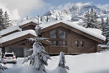 A snow-covered wooden chalet with a parked car, surrounded by pine trees and majestic snowy mountains in the background.