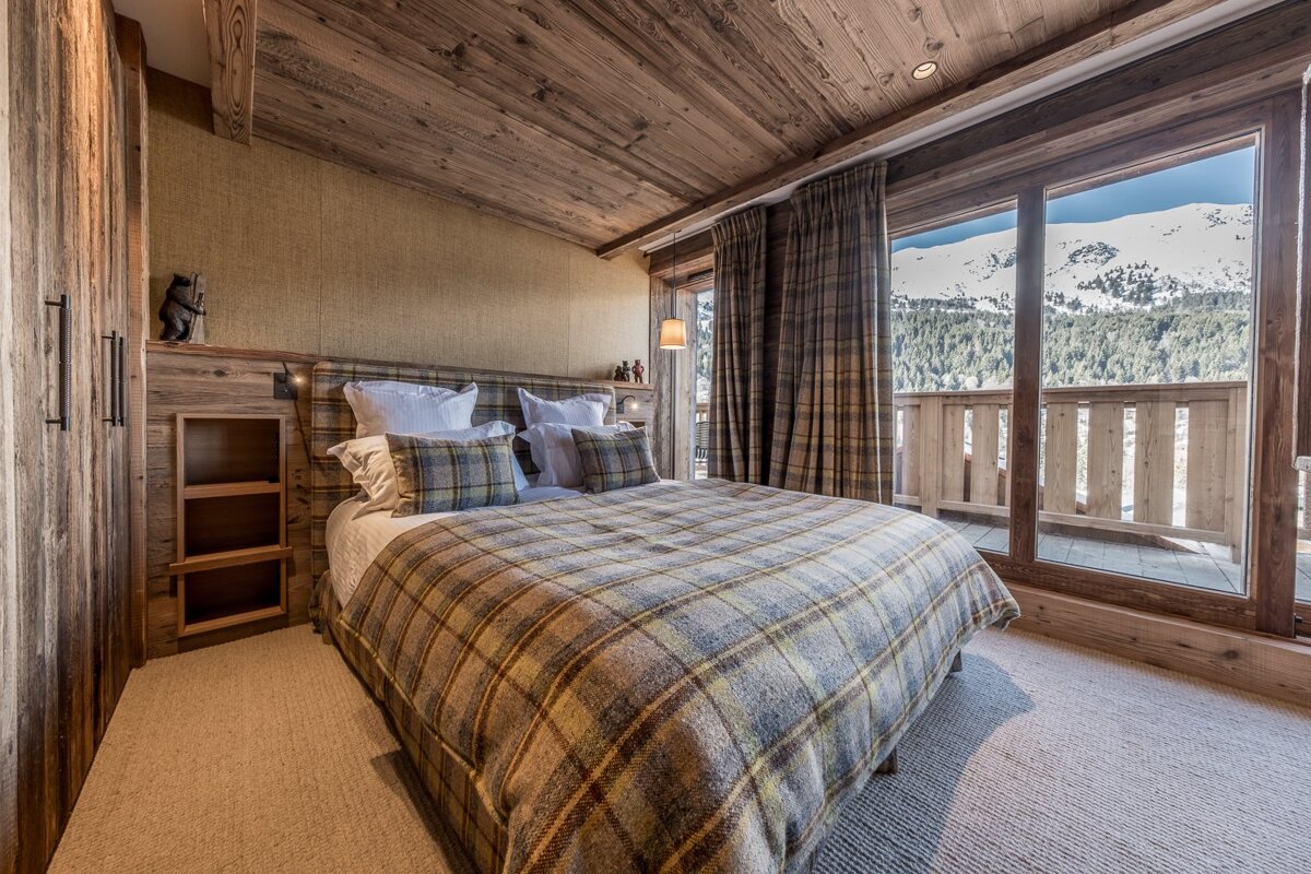 A cozy, wood-paneled bedroom with a plaid bedspread overlooks snowy mountains through a large balcony window.