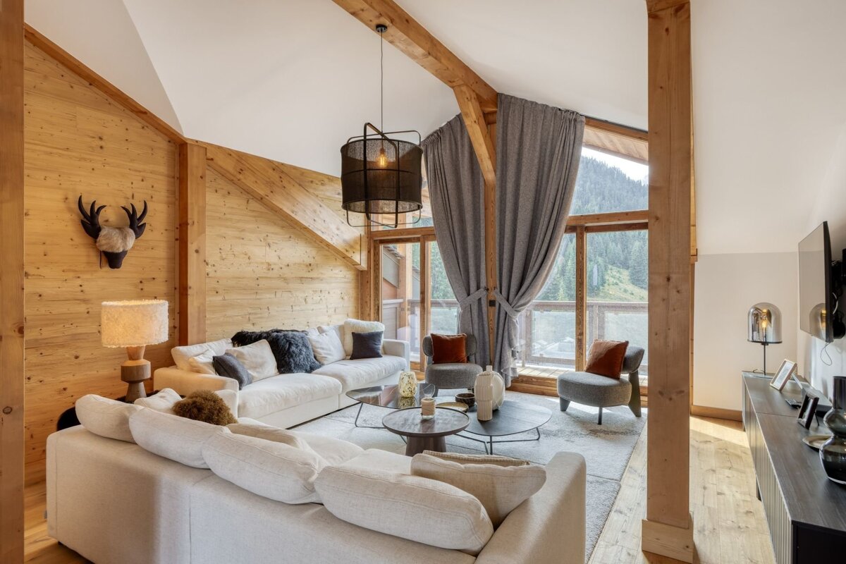 A modern chalet living room featuring warm wood walls, exposed beams, comfy white sofas, and large windows revealing a scenic mountain landscape.