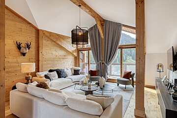 A modern chalet living room featuring warm wood walls, exposed beams, comfy white sofas, and large windows revealing a scenic mountain landscape.