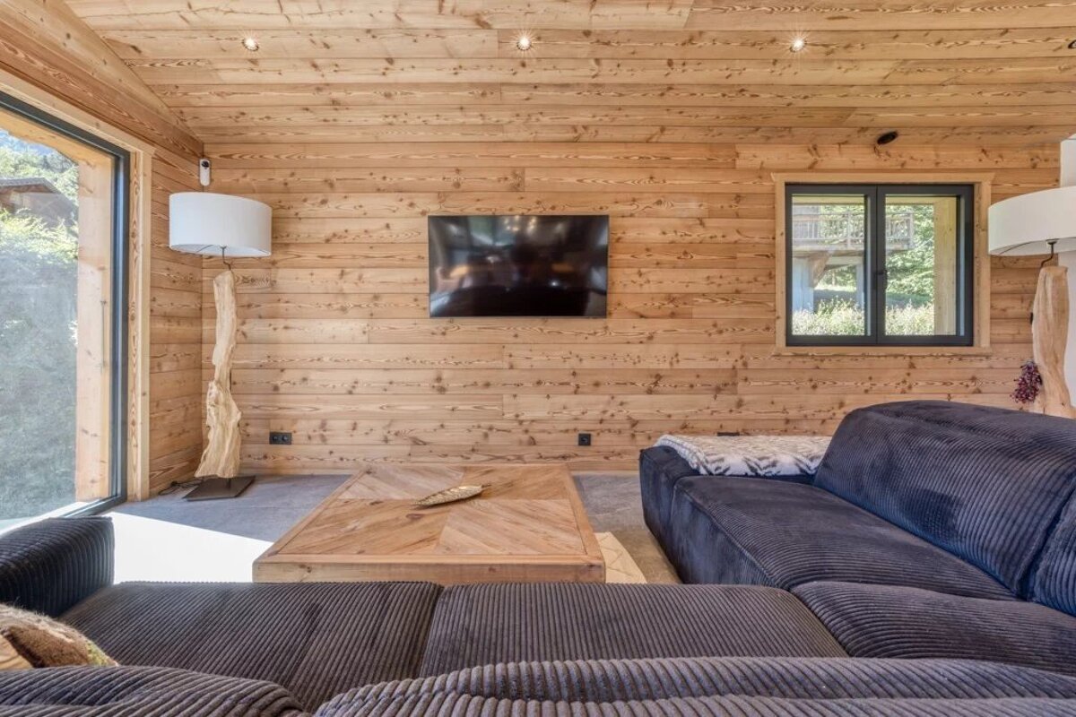 Cozy living room with light wood paneled walls, a dark sectional sofa, a low wooden coffee table, and a wall-mounted TV.