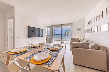 A modern open-plan living/dining room with a set table, wall TV, neutral sofa, and glass doors leading to a balcony with ocean views.
