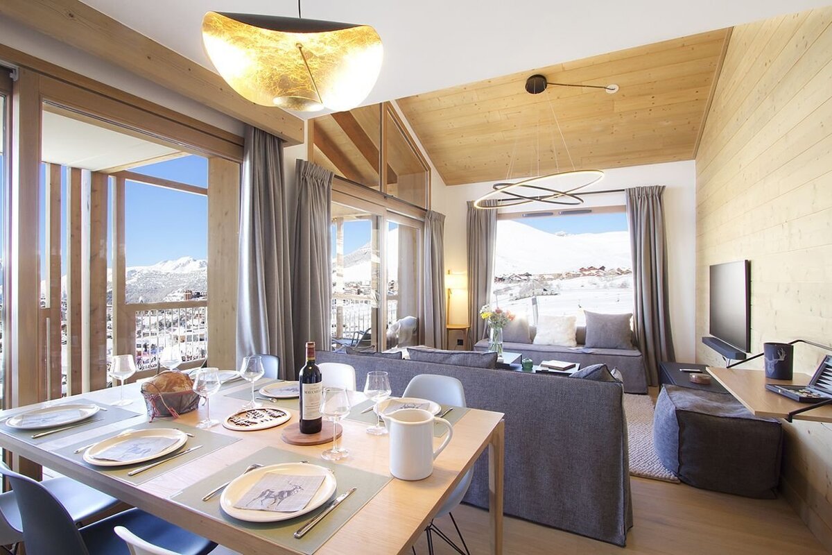 A modern, cozy living and dining room with a prepared table, offering expansive views of snowy mountains through large windows and a balcony.