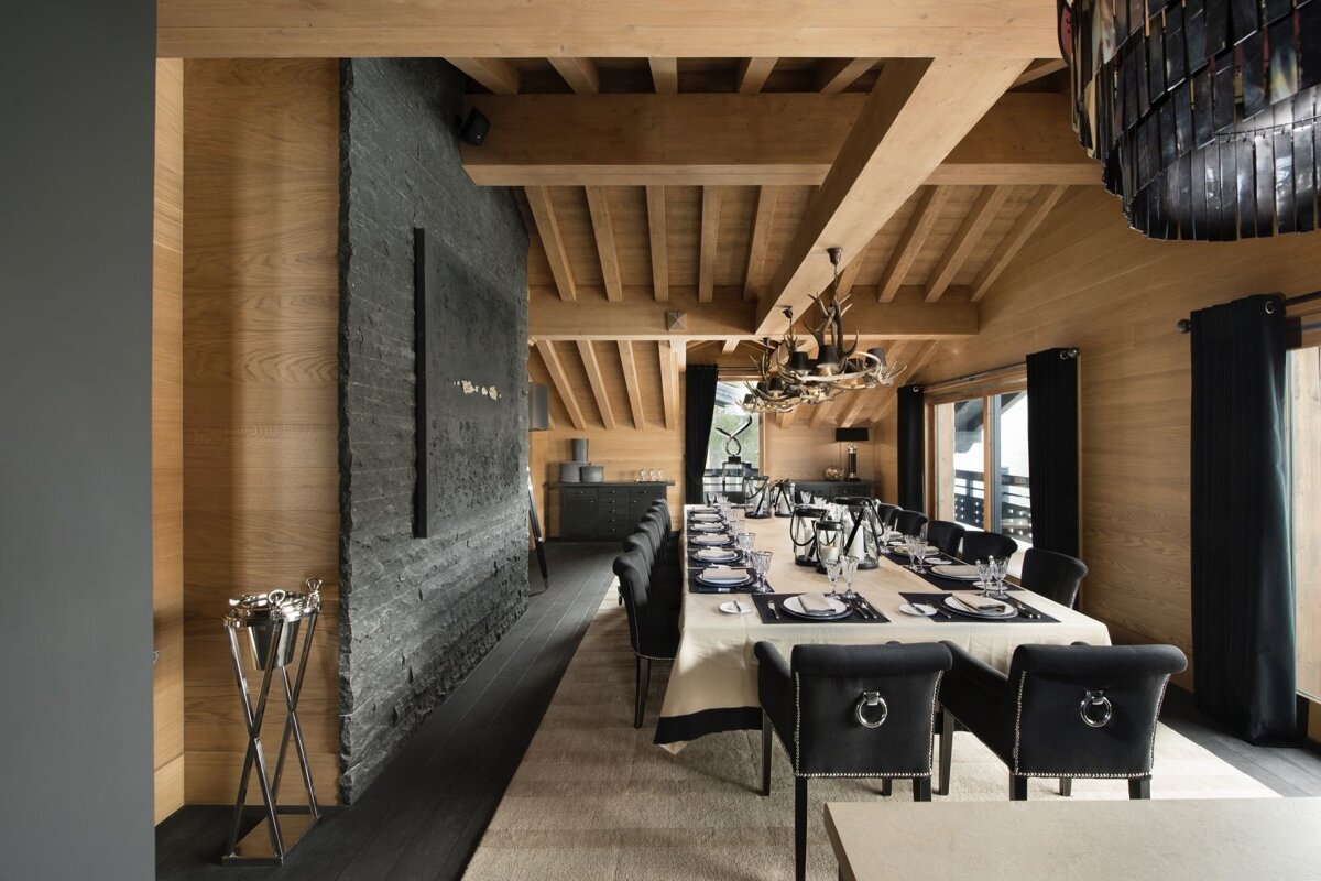 A spacious, modern rustic dining room with a long table set for guests, warm wooden walls/beams, a dark stone accent wall, and unique chandeliers.