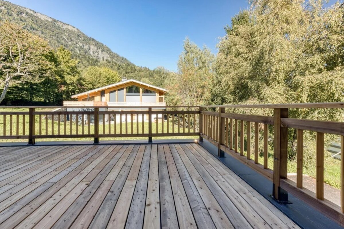 A view from a spacious wooden deck overlooking a modern chalet and green mountains under a clear blue sky.