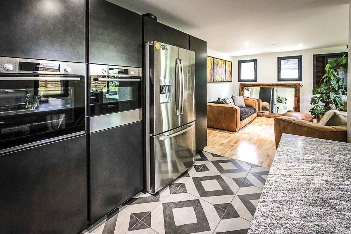 Modern open-plan space featuring a dark kitchen with stainless steel appliances, geometric tile, and a living area with brown sofas and wood floors.