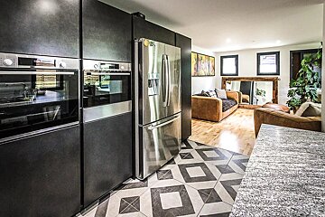 Modern open-plan space featuring a dark kitchen with stainless steel appliances, geometric tile, and a living area with brown sofas and wood floors.