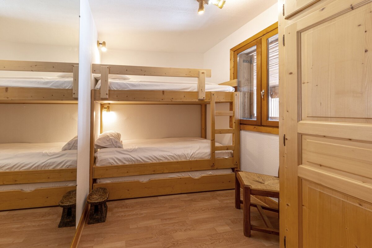 A cozy room features light wood bunk beds with white bedding, a wooden window, and a small chair. A mirror reflects a partial view of another bunk.