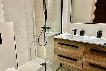 A modern, light-toned bathroom with a walk-in shower, glass partition, and a double vanity featuring wooden cabinets and sleek black fixtures.