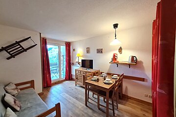 A cozy living/dining area with wooden furniture, a sofa bed, TV, and a balcony with red curtains, adorned with rustic wall decor.