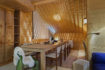 A warm, wood-paneled chalet dining room with a long table, modern chairs, and a slanted ceiling, illuminated by dappled light.