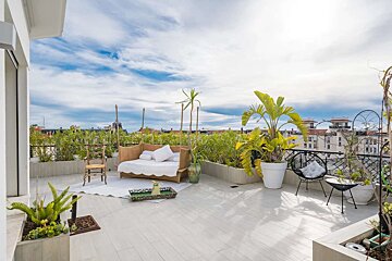A bright, spacious rooftop terrace with wicker outdoor furniture, lush potted plants, and a city skyline view under a partly cloudy sky.