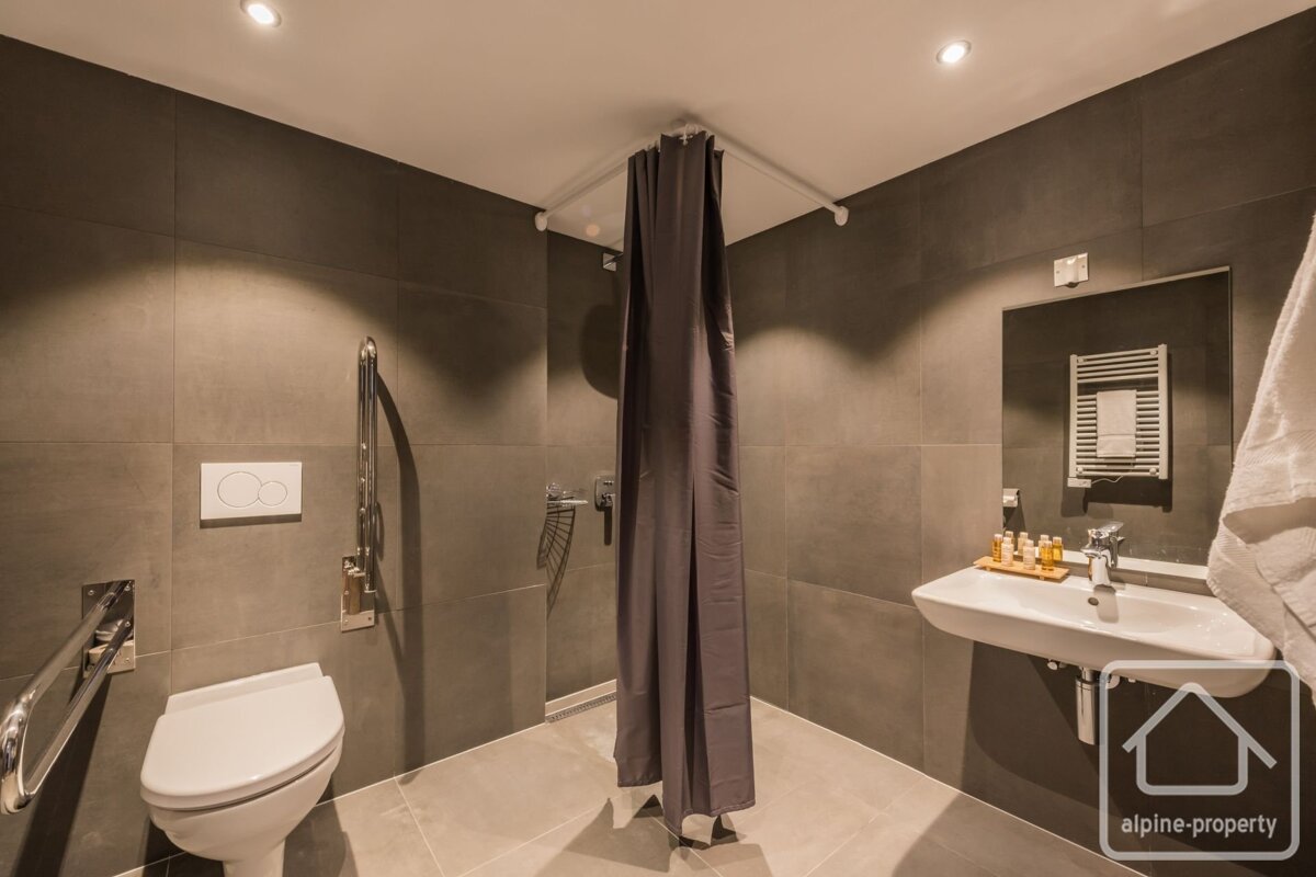 A modern, accessible bathroom with grey tiled walls and floor, featuring a toilet with grab bars, a shower with a grey curtain, and a sink.