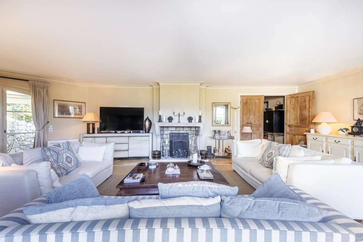 Spacious and bright living room featuring white and blue sofas arranged around a dark wood coffee table, with a fireplace in the background.