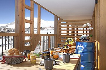 Breakfast on a wooden balcony overlooking a snowy ski resort. Food, drinks, and ski gear are laid out, with a mountain panorama under a clear sky.