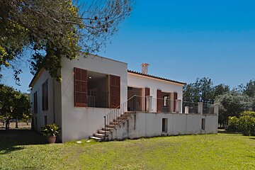 A serene white house with brown shutters and a raised terrace overlooks a lush green lawn, framed by trees under a clear blue sky.