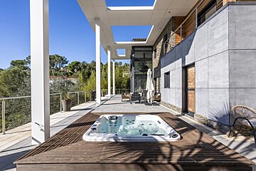 Sunny modern patio with an embedded hot tub on a wooden deck, minimalist columns, contemporary house facade, and a lush green landscape.