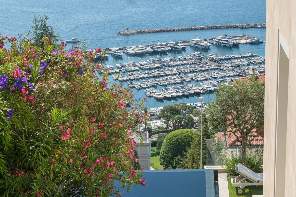 A picturesque high-angle view of a marina filled with yachts, framed by vibrant blooming flowers and a sunny poolside terrace.