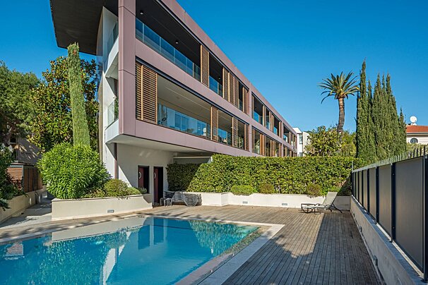 Modern luxury building with a swimming pool, wooden deck, and green hedges under a clear blue sky.