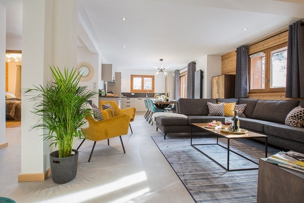Modern open-plan living room with a grey sectional sofa, yellow armchairs, a potted plant, and a dining area in the background.