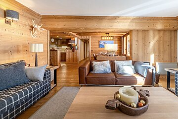Warm, rustic chalet living room with wood paneling, leather and plaid sofas, a deer head, and a dining area, creating a cozy ambiance.