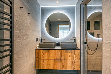 Sleek modern bathroom with dual grey vessel sinks, a natural wood vanity, a large backlit round mirror, and textured grey tiled walls, next to a shower.